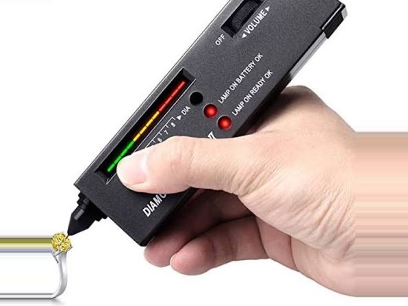 Diamond Tester held in one hand, its probe tip touching a stone set in a ring, with the LED scale lit